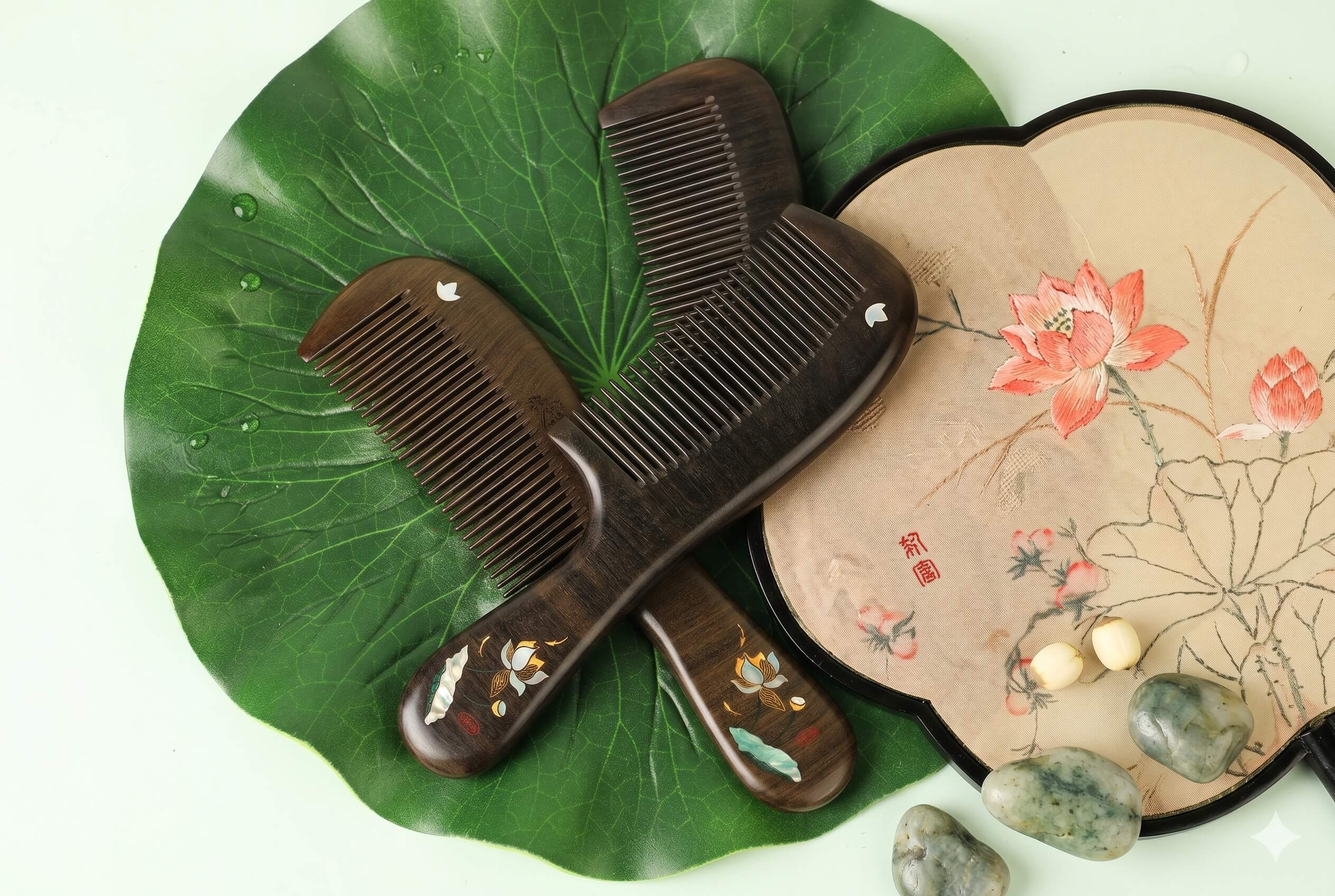 Two wooden combs on a large green leaf with a fan featuring floral design and stones.