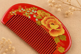 An exquisite traditional Chinese red wooden comb featuring detailed hand-painted golden peony flowers, presented on a textured beige surface with elegant dried blossoms.