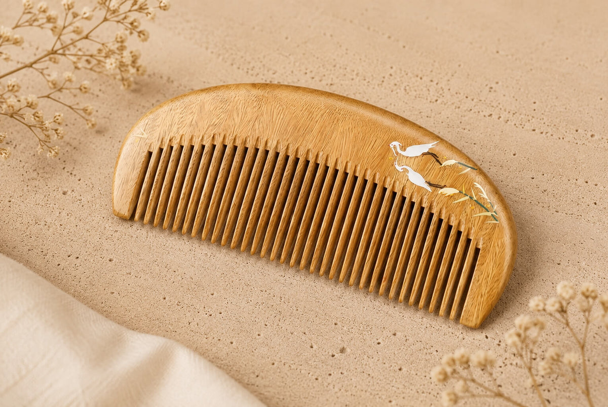 An elegant handcrafted wooden hair comb decorated with fine hand-painted white cranes and reeds, displayed on a textured beige background with dried flowers.