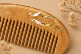 Close-up macro shot of a handcrafted wooden comb featuring exquisite hand-painted details of white cranes and golden leaves against a soft beige background.